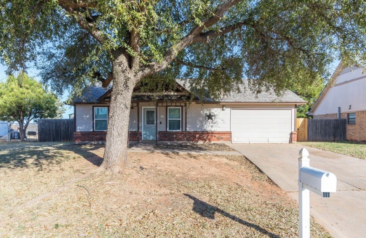 A single-story house with a garage, a mailbox, and a large tree in front.