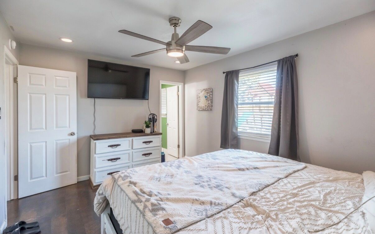 Une chambre à coucher comprenant un lit, un ventilateur de plafond, une commode, un téléviseur fixé au mur et une fenêtre avec des rideaux.