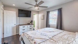 Ein Schlafzimmer mit einem Bett, einem Deckenventilator, einem Schminktisch, einem an der Wand befestigten Fernseher und einem Fenster mit Vorhängen.