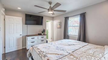 Une chambre à coucher comprenant un lit, un ventilateur de plafond, une commode, un téléviseur fixé au mur et une fenêtre avec des rideaux.