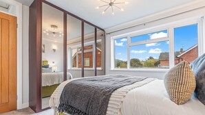 Une chambre avec une grande fenêtre, un lit recouvert d’une couverture grise et un miroir qui reflète la pièce.