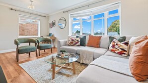 Un salon moderne avec un canapé, des fauteuils, une table basse en verre et une vue sur l’extérieur grâce à de grandes fenêtres.
