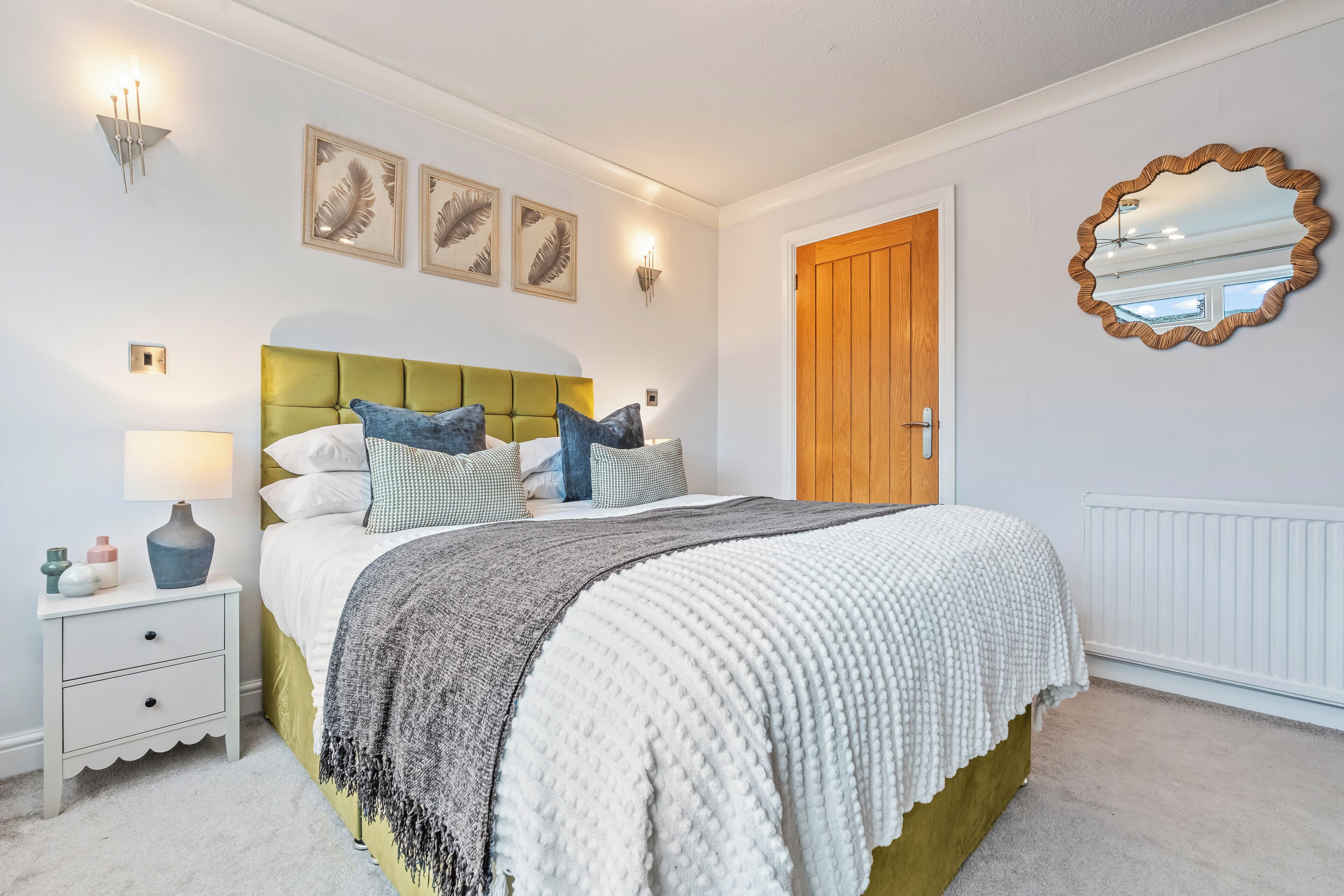 Une chambre à coucher avec une tête de lit recouverte d’un tissu vert, une couvre-lit blanche et un plaid gris. Des appliques murales et un miroir sont fixés au mur au-dessus du lit.