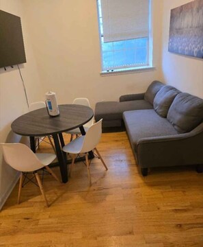 Ein Wohnzimmer mit rundem Tisch, zwei Stühlen, einem grauen Sofa und einem Fenster mit Jalousien.