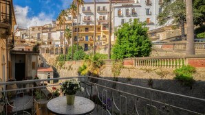 A balcony with a round table and chairs, overlooking a street with buildings and a stone wall. - La Colombaia Bed and Rooms (Belvedere Marittimo)