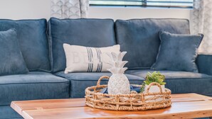 A living room with a blue sofa, a wooden coffee table, a woven basket with a pineapple-shaped vase and a plant. - Sleeps 12! Ocean Views! 4 BD/4BA! 2 Renovated Condos! (Kihei)