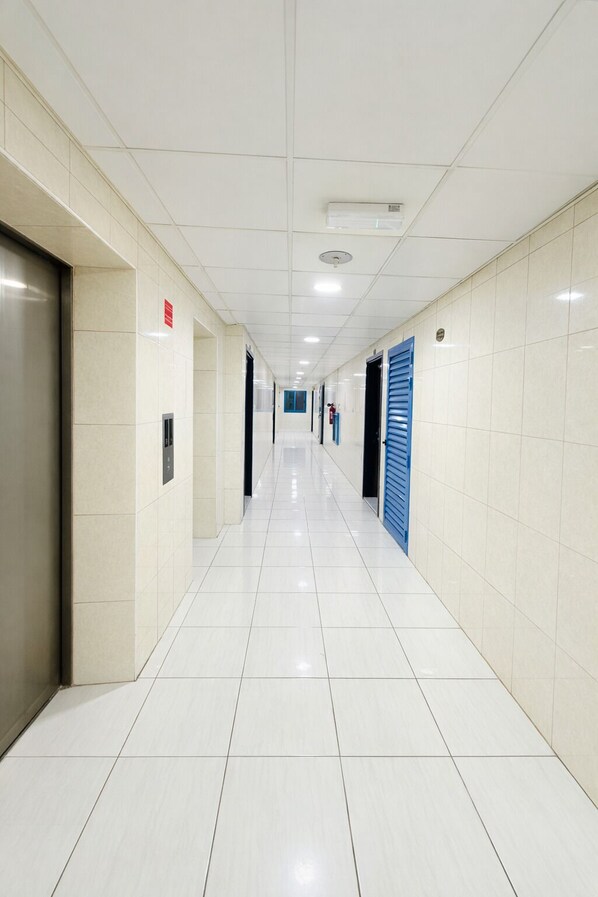 A clean, well-lit hallway with white tiled floors and walls, featuring doors and a blue shutter on the right. - TIDY Premium Hostel (Sharjah)