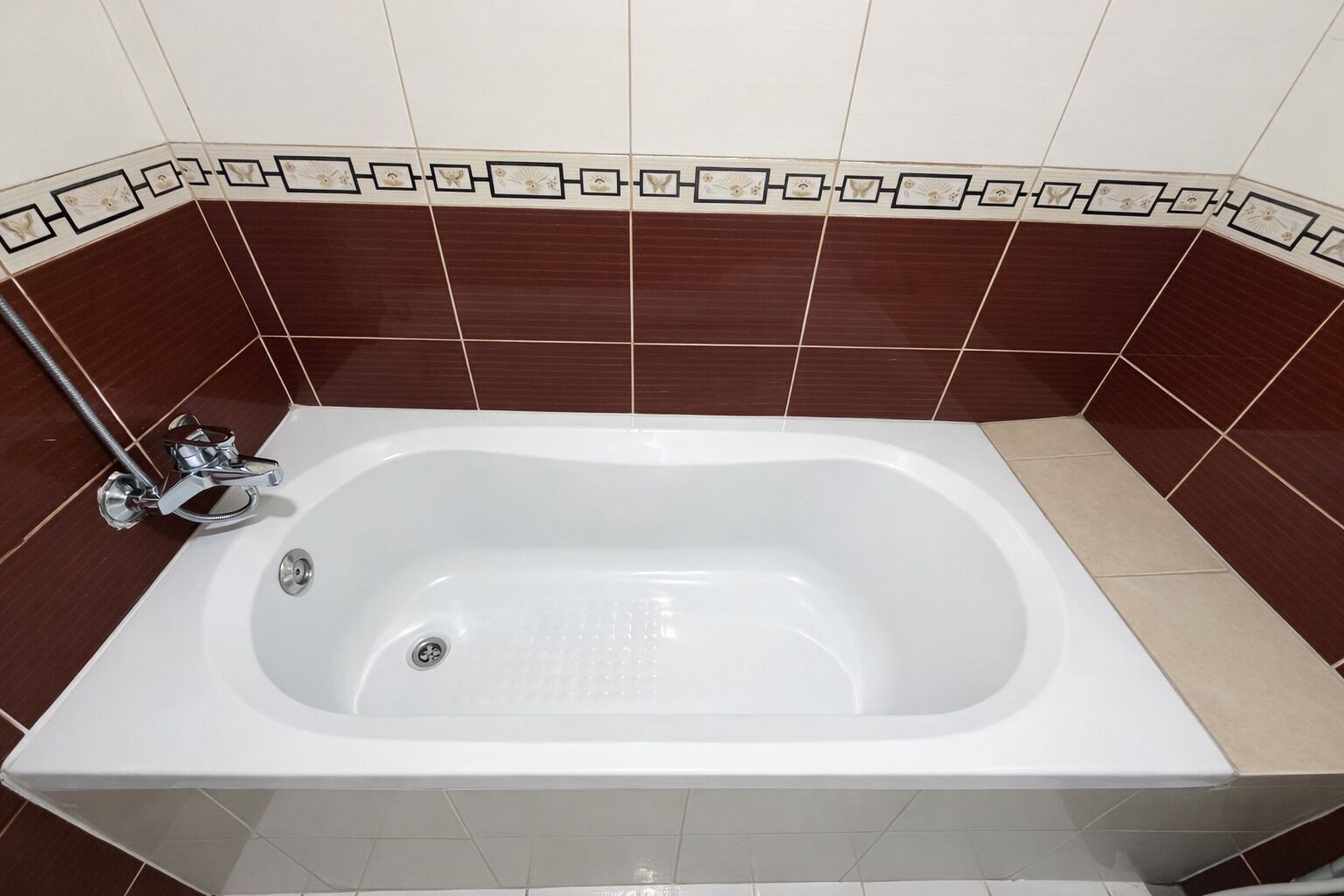 Une baignoire rectangulaire dotée d’un robinet et d’un drain en chrome, placée devant un mur recouvert de carreaux bruns et beiges, le tout agrémenté d’une bordure décorative.
