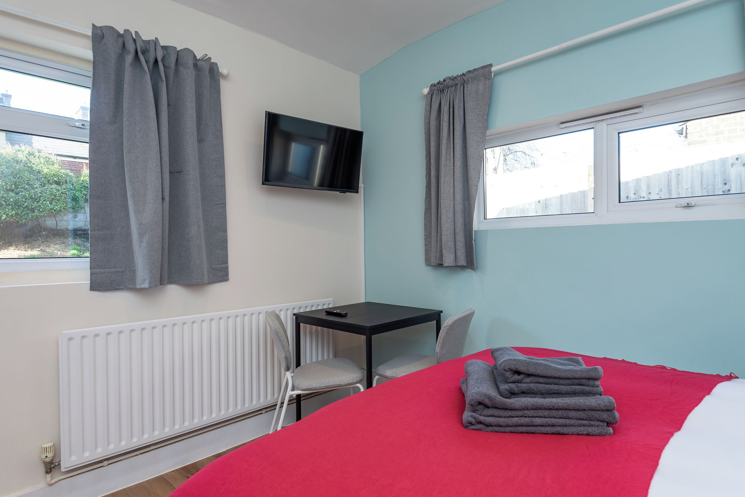 Quarto com cama, escrivaninha, cadeira, televisão e janela com cortinas.