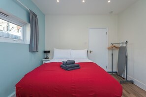 A bedroom with a bed, a nightstand, a window with curtains, a door, and a coat rack with a coat. - Modern Studio With Parking And Private Garden (Greater London)