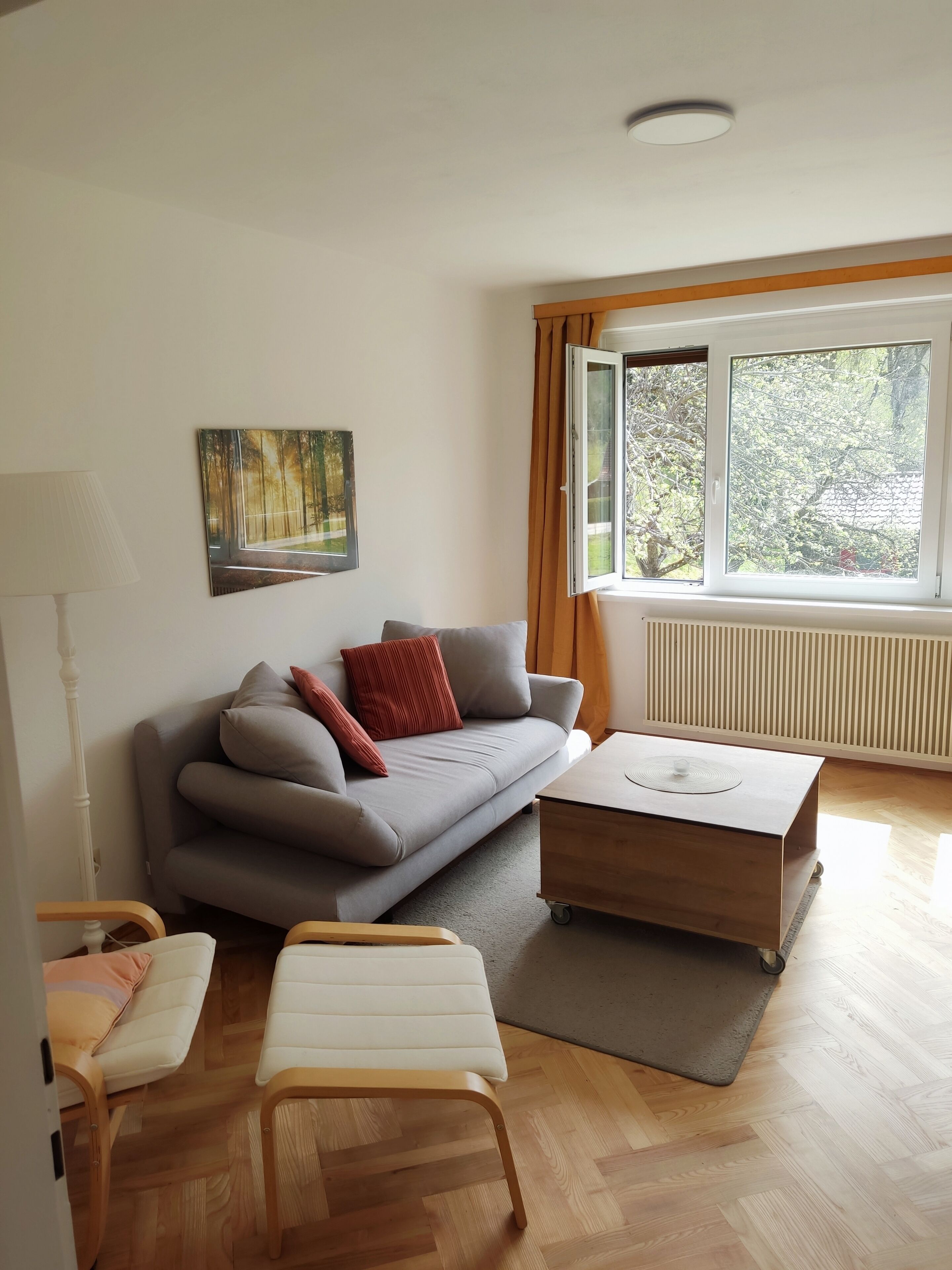 Una sala de estar moderna con un sofá gris, una mesa de centro de madera y un sillón blanco. Hay un cuadro en la pared y se ve una ventana con cortinas.