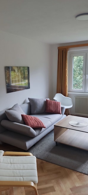 A modern living room with a grey sofa, a wooden coffee table, and a white armchair. A painting of a forest is on the wall, and a window with curtains is visible.