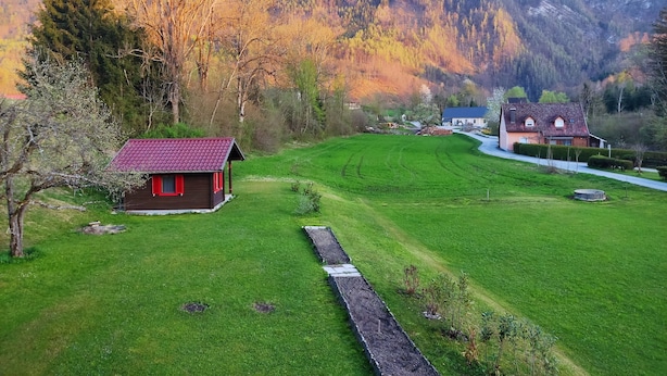 A rural landscape with a small red-roofed cabin and a larger house with a blue roof, surrounded by lush green fields and mountains in the background. - Variety of lakes: Klopeiner See, Gösselsdorfer See, Turner See. (Sittersdorf)
