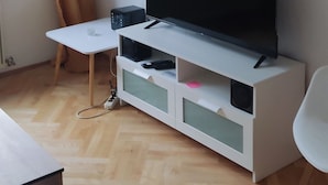 A living room with a flat-screen TV on a white entertainment unit, a small white table, and a window with curtains. - Variety of lakes: Klopeiner See, Gösselsdorfer See, Turner See. (Sittersdorf)