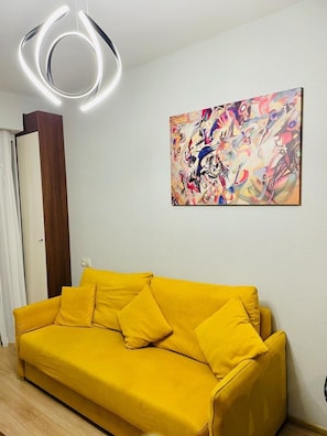 A yellow sofa with pillows, a modern light fixture, and a colorful abstract painting on the wall. - Crystal Residence – Balcony with Ski Slope View (Bakuriani)