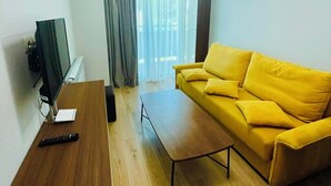 Ein modernes Wohnzimmer mit einem gelben Sofa, einem Holztisch, einem Fernseher auf einem Schrank und einem großen Fenster mit Vorhängen.