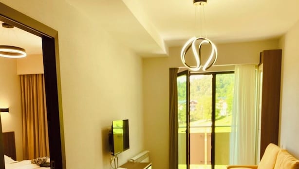 Ein modernes Hotelzimmer mit Holzboden, einem Bett, einem Schreibtisch mit Fernseher, einem Sofa und einem Balkon mit Vorhängen.