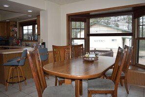 A dining area with a wooden table and chairs, a kitchen in the background, and a view through large windows. - Woodland Retreat with 40 acre and 4 Bedrooms Perfect for Families & Dogs (Mauston)