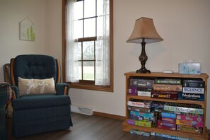 A room with a teal armchair, a wooden bookshelf with board games, a window with sheer curtains, and a table lamp. - Woodland Retreat with 40 acre and 4 Bedrooms Perfect for Families & Dogs (Mauston)