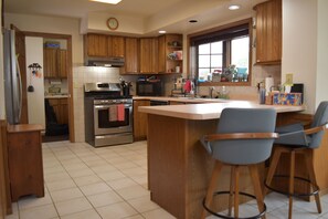 A kitchen with wooden cabinets, a stainless steel refrigerator, an oven, a microwave, and a sink. There are two bar stools and a window with a view of the outside. - Woodland Retreat with 40 acre and 4 Bedrooms Perfect for Families & Dogs (Mauston)