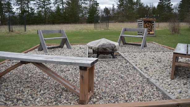 A wooden deck with two benches and a table, surrounded by gravel and a grassy area with trees in the background.
