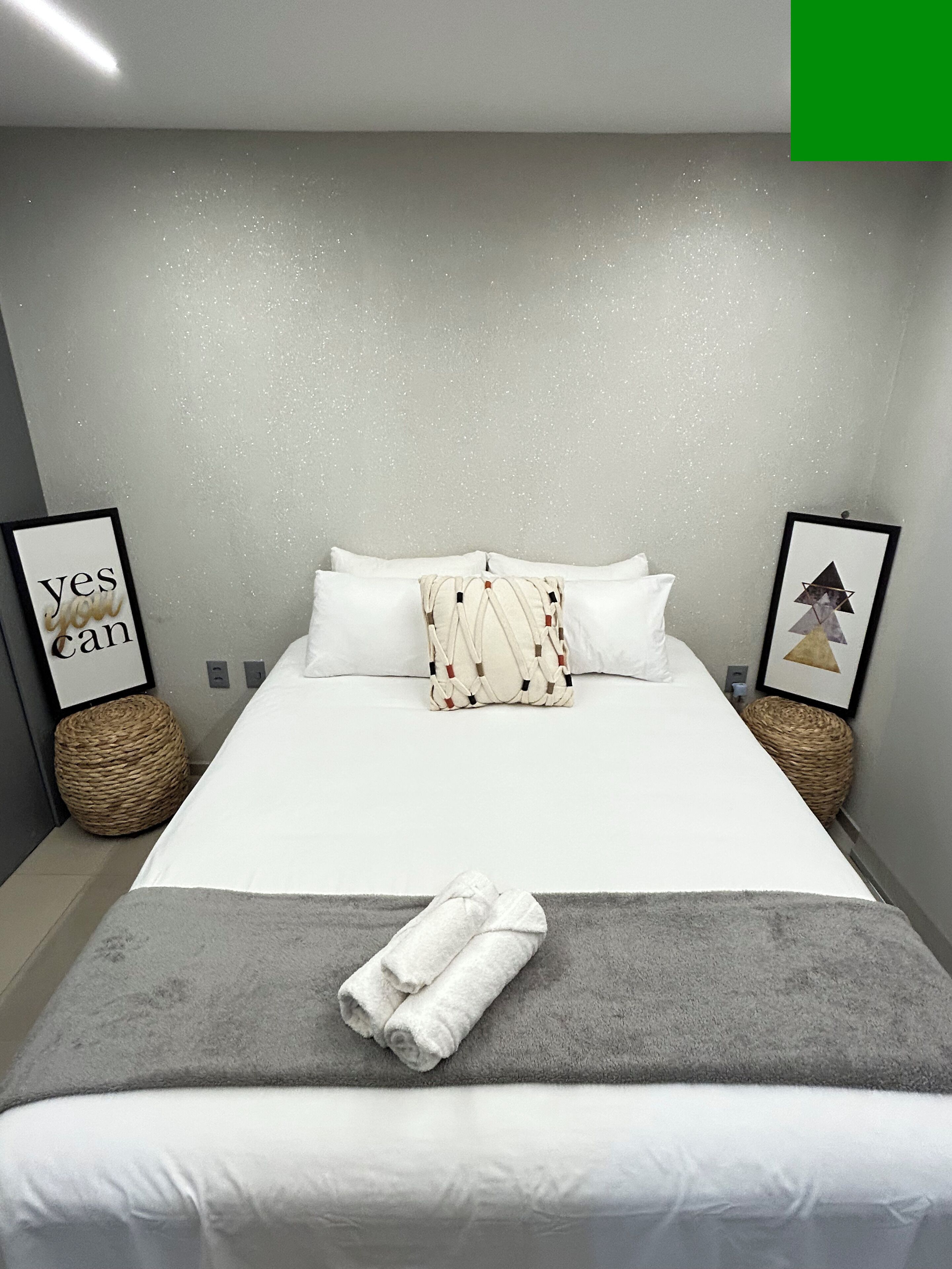 Un lit bien fait, avec des draps blancs, un plaid gris et des serviettes pliées. Deux paniers en osier sont posés sur le sol, à côté du lit.