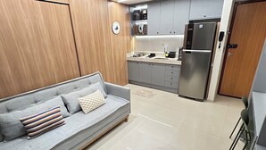 A modern kitchen with a grey sofa, wooden cabinets, and a stainless steel refrigerator. - Melhores Flats - Espaçoso pé na areia (João Pessoa)