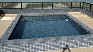 A rectangular pool with tiled edges, surrounded by a glass railing, overlooking a calm sea under a partly cloudy sky. - Melhores Flats - Espaçoso pé na areia (João Pessoa)