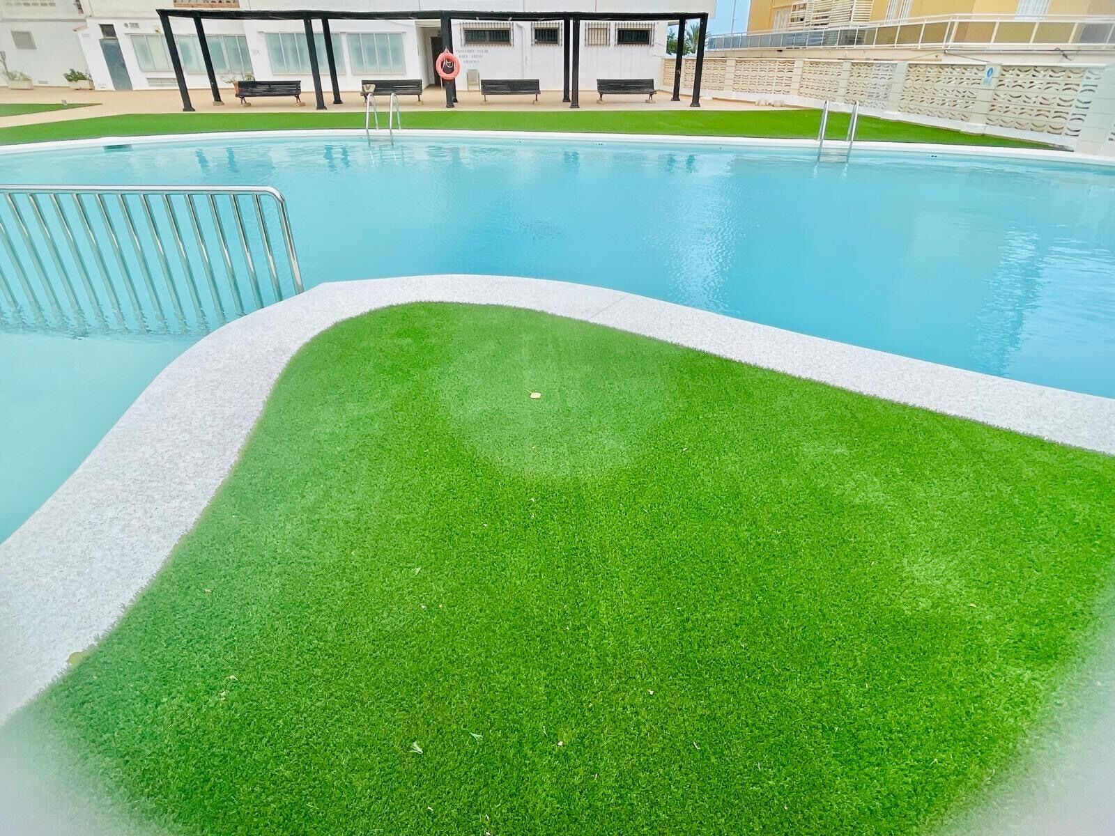 En swimmingpool med en græsplæne i forgrunden og et overdækket opholdsområde i baggrunden.