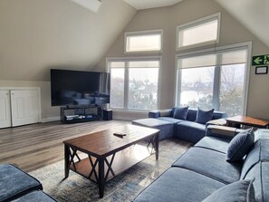 A modern living room with a sectional sofa, a wooden coffee table, a flat-screen TV, and large windows offering a view of the outdoors.