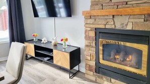 A modern living room with a flat-screen TV mounted on the wall, a black entertainment unit with decorative vases, a stone fireplace with a glass front, and a light-colored chair. - Private Ski Chalet Hot Tub & Mountain Views (The Blue Mountains)