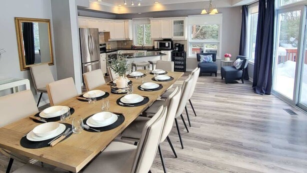 A dining area with a long wooden table set for six, surrounded by chairs. There is a kitchen in the background with modern appliances. - Private Ski Chalet Hot Tub & Mountain Views (The Blue Mountains)
