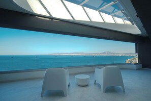 Ein modernes Zimmer mit einem großen Panoramafenster mit Meerblick, zwei weißen Stühlen und einem runden Tisch.