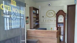 A hotel reception area with a wooden reception desk, a grey sofa, and a wall-mounted clock. - HOTEL NOVAES (Castanhal)