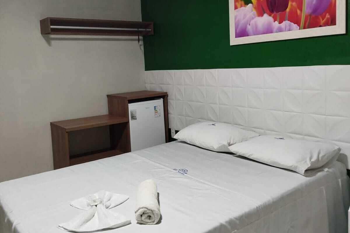 A hotel room with a bed, a small desk, a refrigerator, and a green wall with a flower painting.