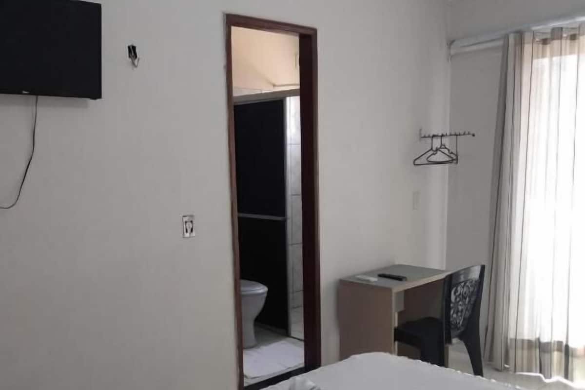 A hotel room with a bed, a desk, a chair, a TV mounted on the wall, and a bathroom visible through an open door.