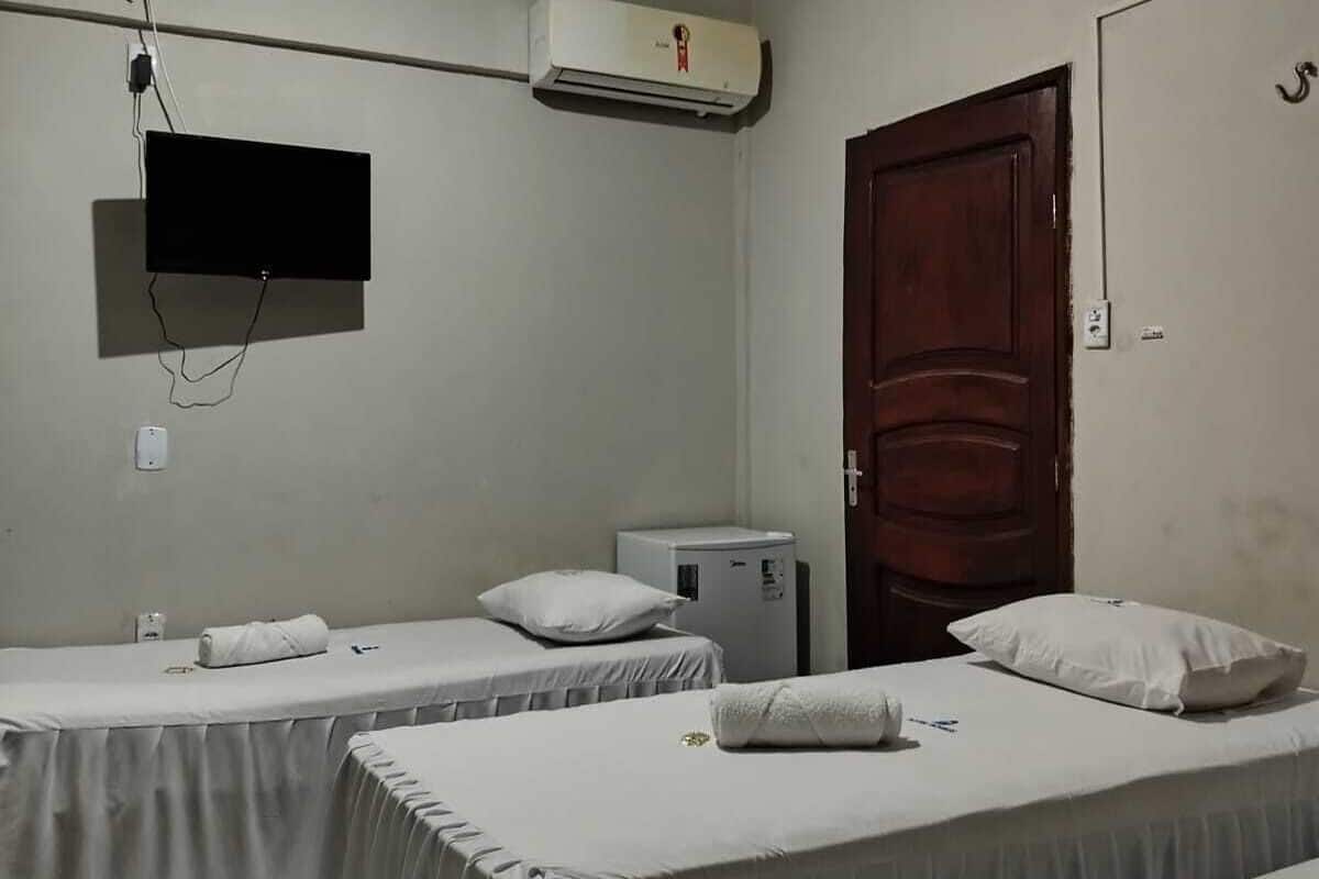A room with two single beds, a TV mounted on the wall, an air conditioner, a small refrigerator, and a wooden door.