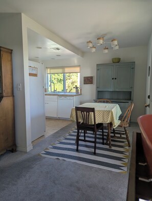 Un coin repas avec une table dressée pour deux personnes, une cuisine en arrière-plan et une fenêtre laissant entrer la lumière naturelle.