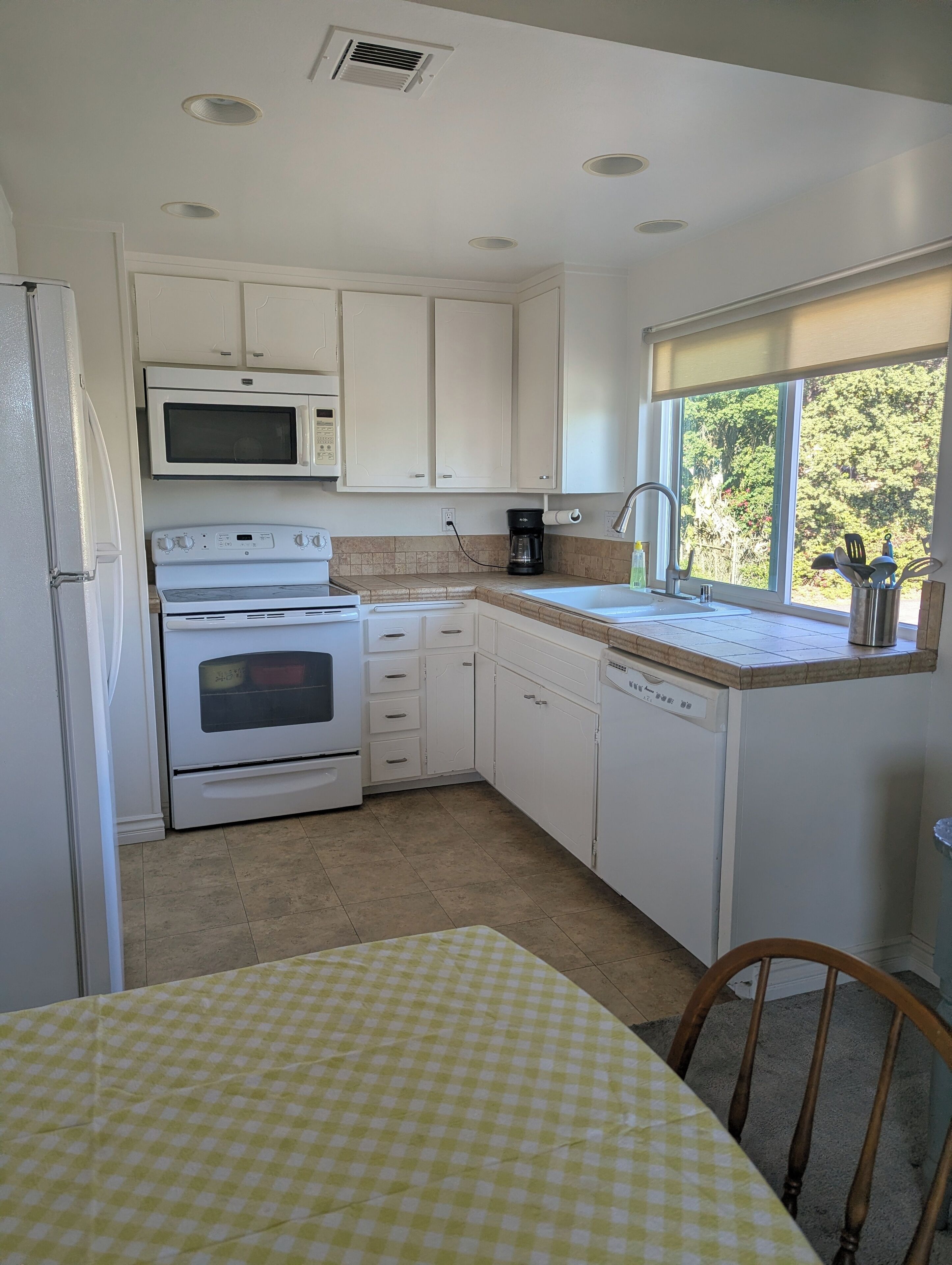 A compact kitchen with white cabinets, a microwave, a stove, a dishwasher, and a window with a view of trees.