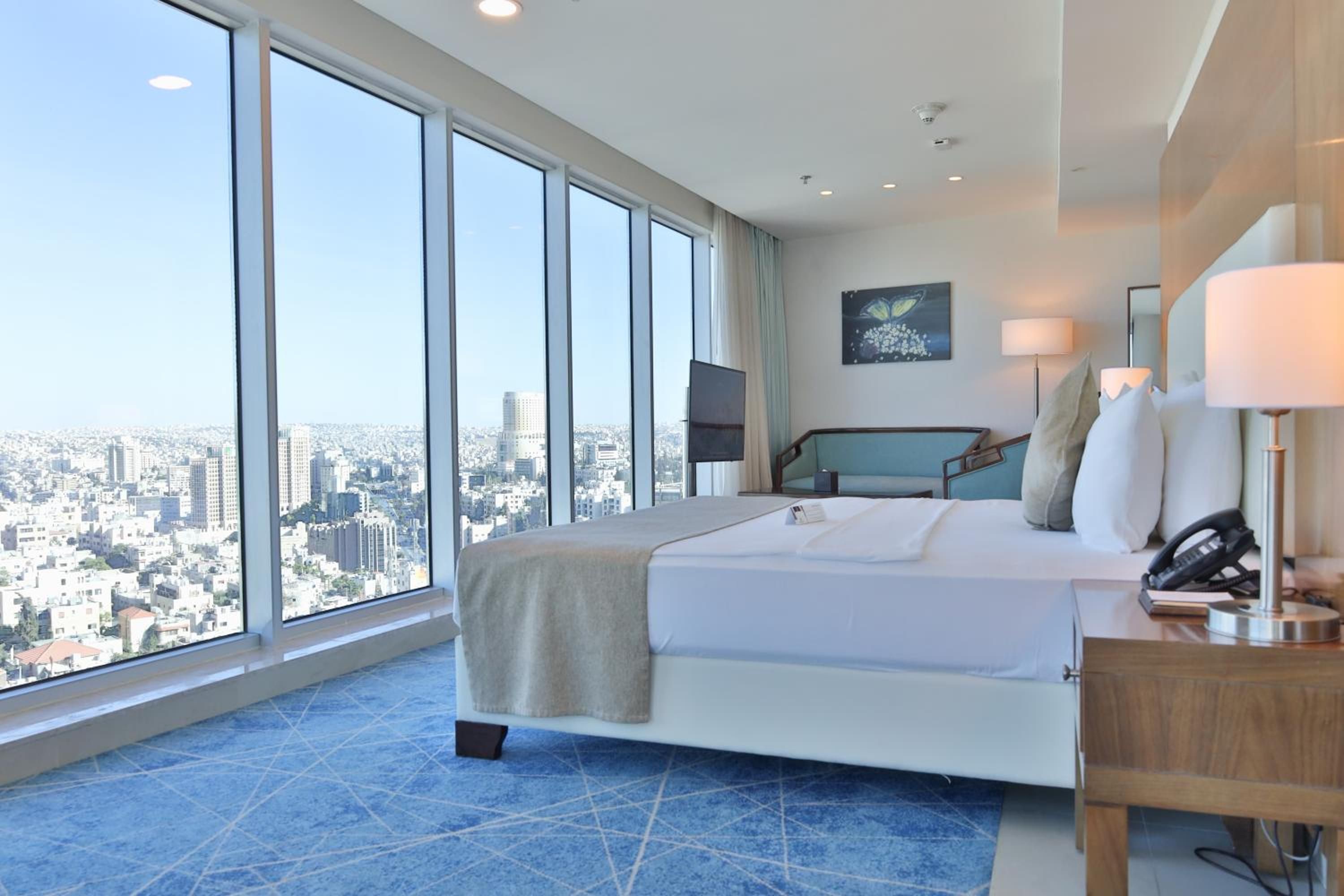 A modern hotel room with a large bed, a desk, and a view of a cityscape through floor-to-ceiling windows.