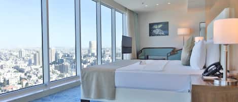 A modern hotel room with a large bed, a desk, and a view of a cityscape through floor-to-ceiling windows.