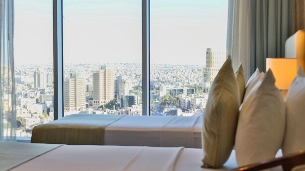 Ein Hotelzimmer mit großem Fenster, einem Bett mit Kissen und Blick auf eine Stadtlandschaft.