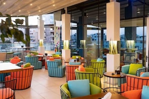 Un espacio interior moderno con asientos y mesas coloridos, ventanas amplias y vistas de edificios.