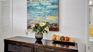 Une pièce comprenant une commode en bois, un tableau représentant un paysage bleu et un vase rempli de fleurs et de fruits, posé sur la commode.