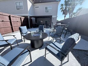 En udendørs patio med et rundt bord og stole, en bålplads og et overdækket siddeområde.