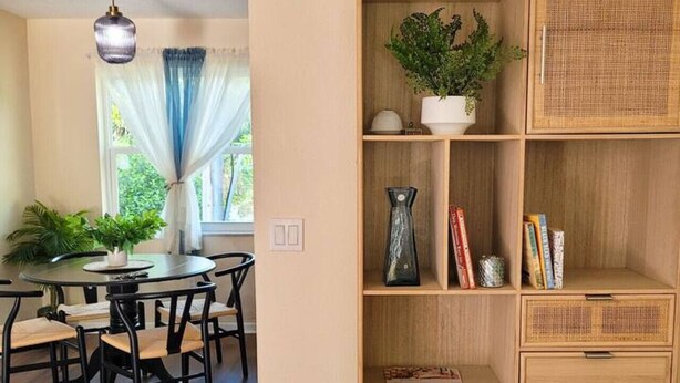 Uma sala de jantar com uma mesa redonda e cadeiras, uma estante com prateleiras de vime e uma janela com cortinas.