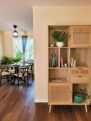 A dining area with a round table and chairs, a bookshelf with wicker shelves, and a window with curtains. - St. Pete Boho: Central 2-BR, heated pool, pets ok! (Pinellas Park)