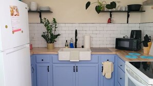 Uma cozinha com armários azuis, geladeira branca, pia branca, micro-ondas e fogão. Há uma planta pendurada, uma estante com louças e uma luminária de teto trançada.