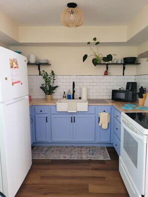 Een keuken met blauwe kastjes, een witte koelkast, een wit aanrecht, een magnetron en een fornuis. Er hangt een plant, een plank met servies en een geweven plafondlamp.