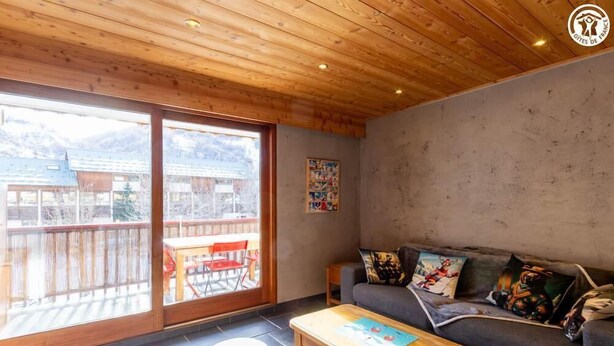 Een gezellige woonkamer met een houten plafond, een grijze bank met kleurrijke kussens, een houten salontafel en een grote schuifdeur naar een terras.